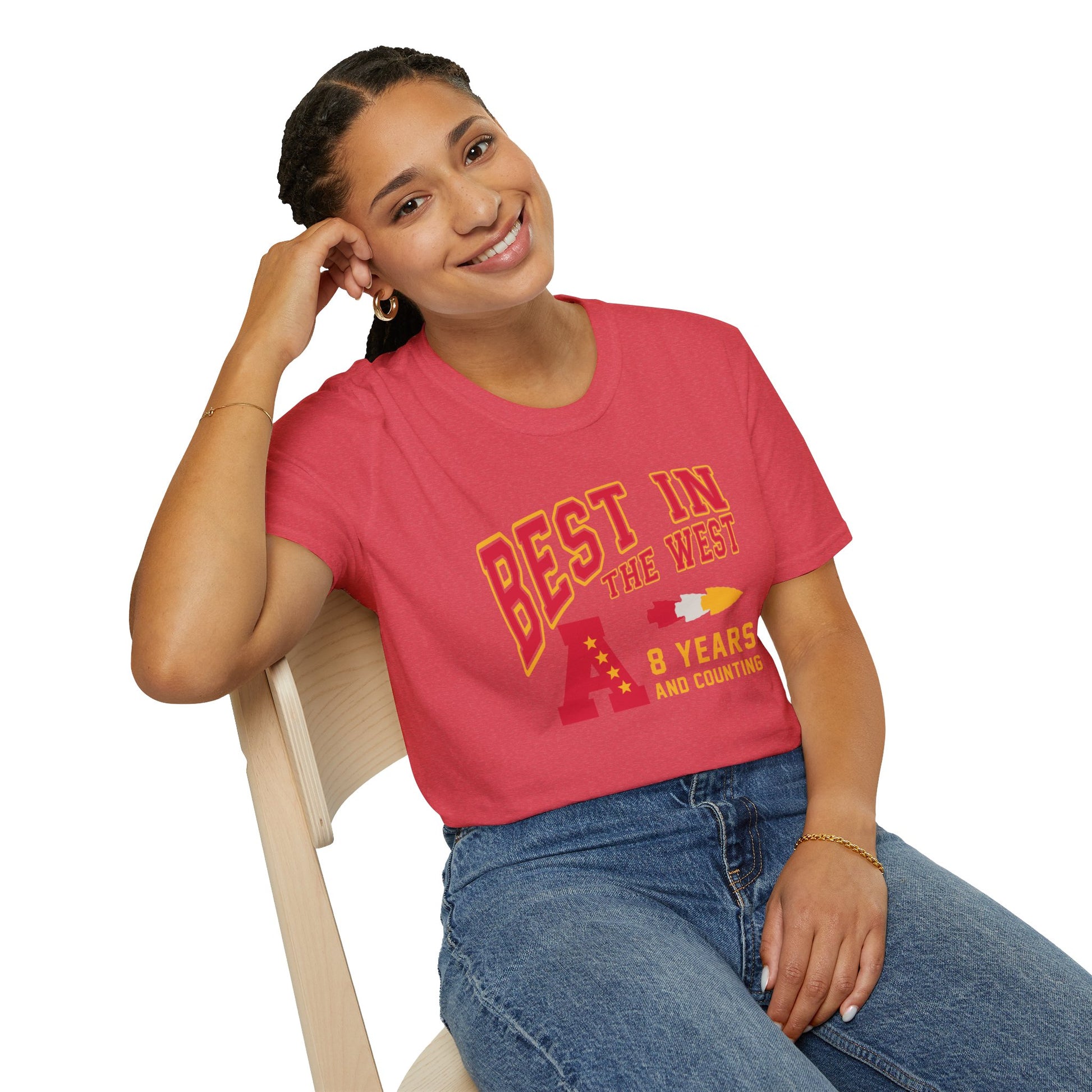 A smiling person wearing a red t-shirt that reads "Best In The West - Kansas City Chiefs Inspired T-Shirt" by KC Inspirations sits on a light-colored wooden chair. The person, appearing to celebrate another AFC West Division Title like the Kansas City Chiefs, has braided hair and rests their left elbow on the chair's back while their left hand touches the side of their head.