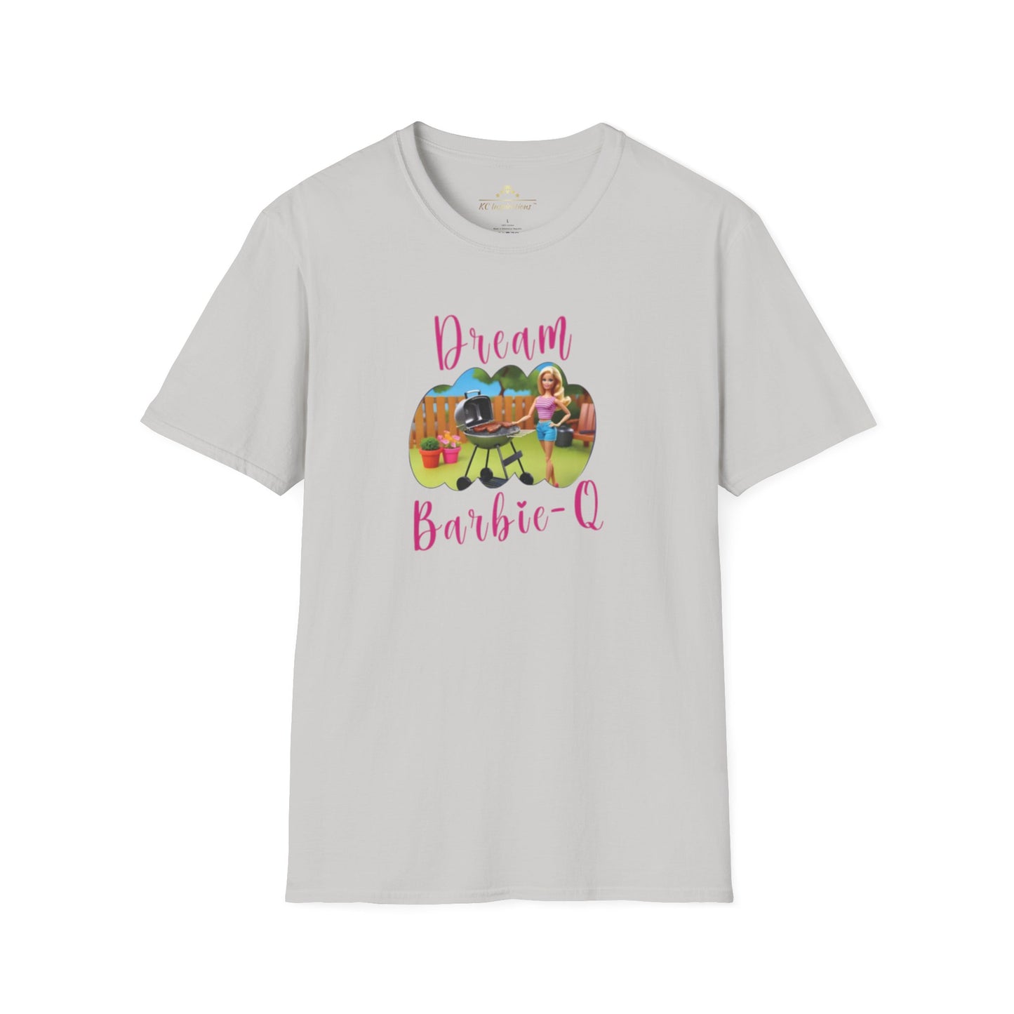 A beige T-shirt with speckles featuring a crisp graphic design of two dolls barbecuing in a backyard. The text "Dream Barbie-Q" is written in pink cursive above and below the image, making this Dream Barbie Q - K.C. BBQ Inspired T-Shirt by KC Inspirations perfect for fans of Kansas City BBQ.