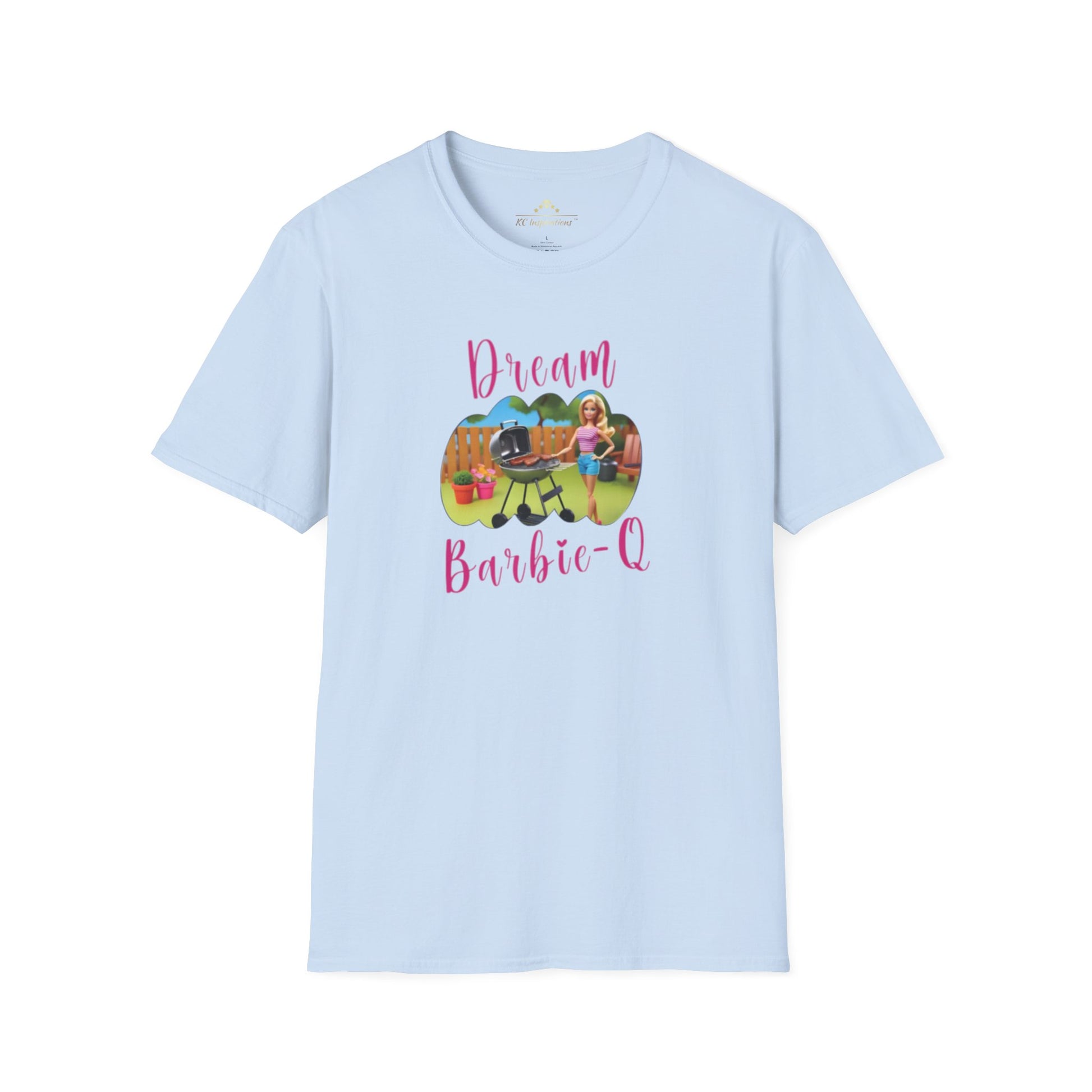 A beige T-shirt with speckles featuring a crisp graphic design of two dolls barbecuing in a backyard. The text "Dream Barbie-Q" is written in pink cursive above and below the image, making this Dream Barbie Q - K.C. BBQ Inspired T-Shirt by KC Inspirations perfect for fans of Kansas City BBQ.