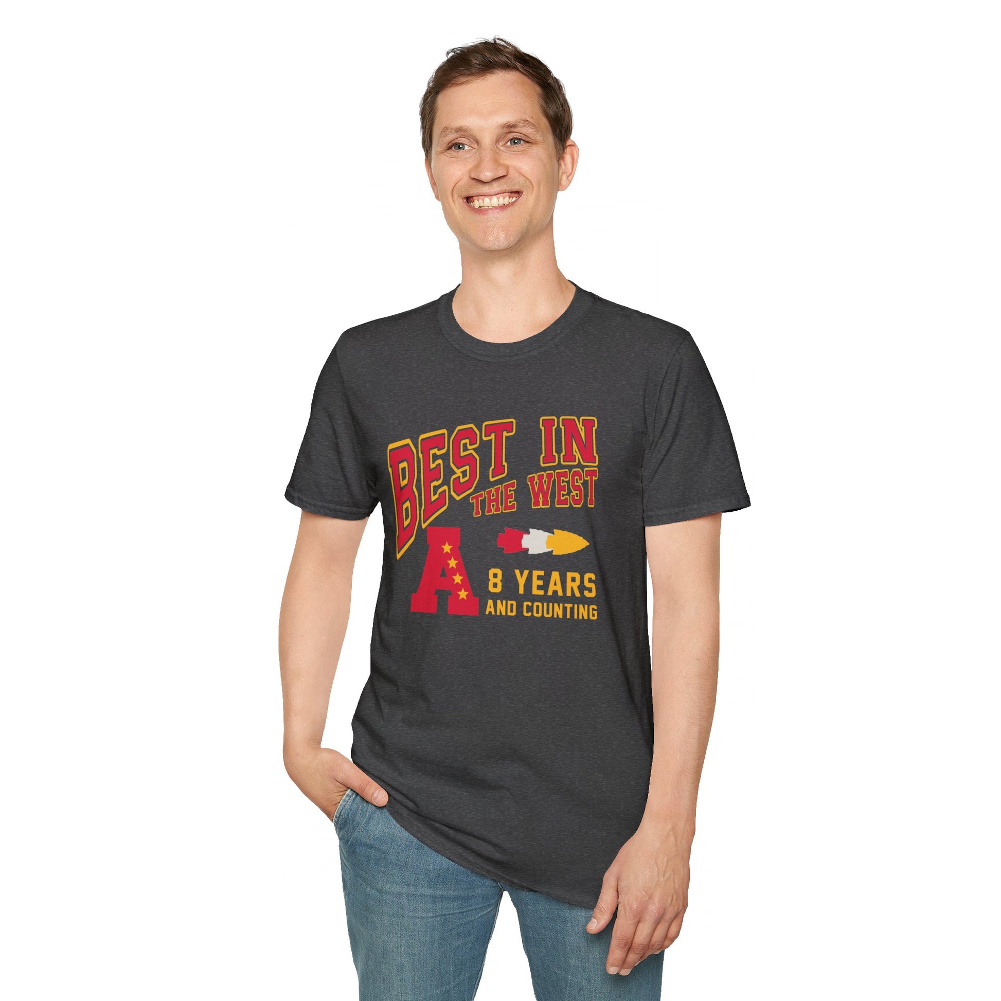 A person stands smiling, hands in pockets, wearing a gray "Best In The West - Kansas City Chiefs Inspired T-Shirt" by KC Inspirations with red and yellow text that reads, "Best in the West, 8 Years and Counting," alongside graphics of a letter "A" and a rocket, proudly representing the Kansas City Chiefs' AFC West Division Title.