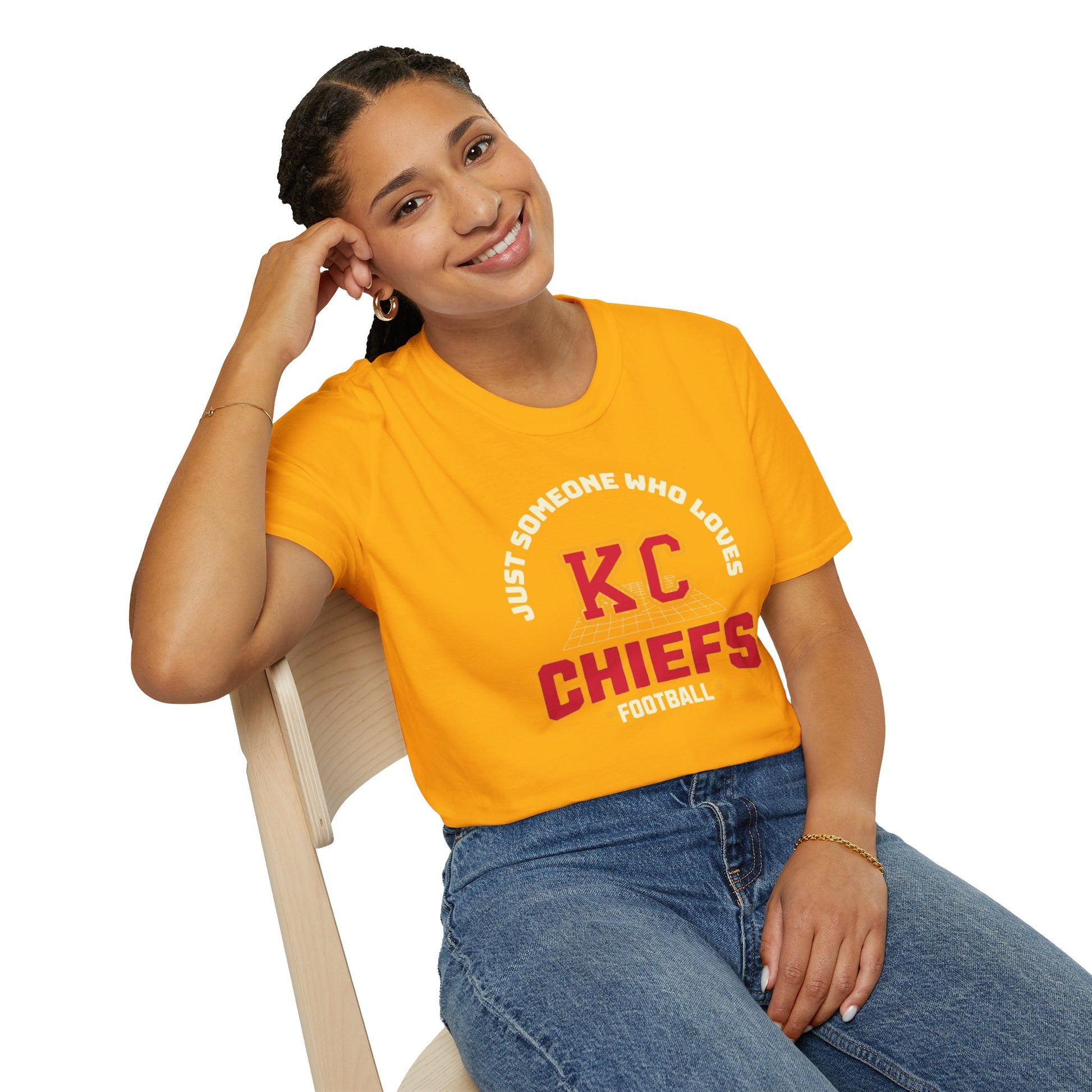 A smiling person with braids sits on a chair, resting their head on one hand. They proudly wear an orange T-shirt from KC Inspirations, titled "Just Someone Who Loves - Kansas City Chiefs Inspired T-Shirt," showcasing their Chiefs pride, along with blue jeans and a gold bracelet.