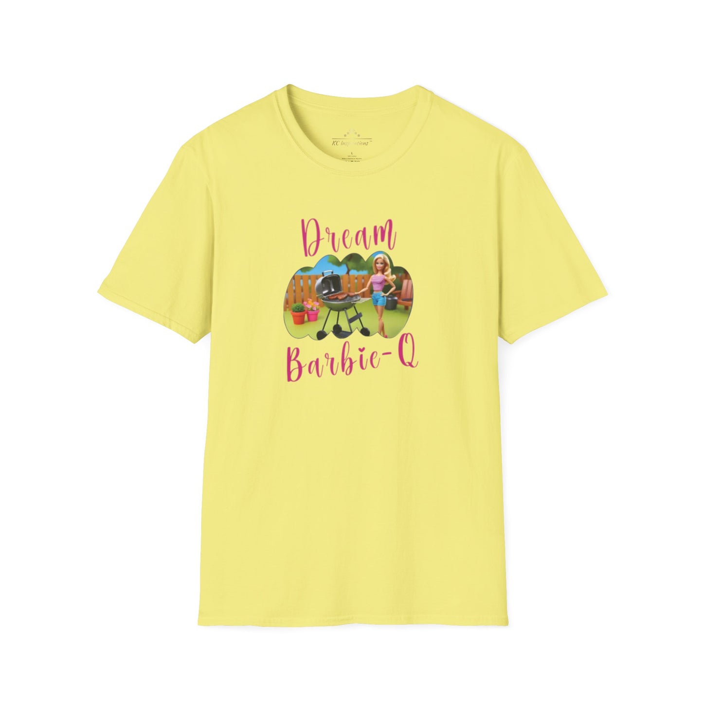 A beige T-shirt with speckles featuring a crisp graphic design of two dolls barbecuing in a backyard. The text "Dream Barbie-Q" is written in pink cursive above and below the image, making this Dream Barbie Q - K.C. BBQ Inspired T-Shirt by KC Inspirations perfect for fans of Kansas City BBQ.