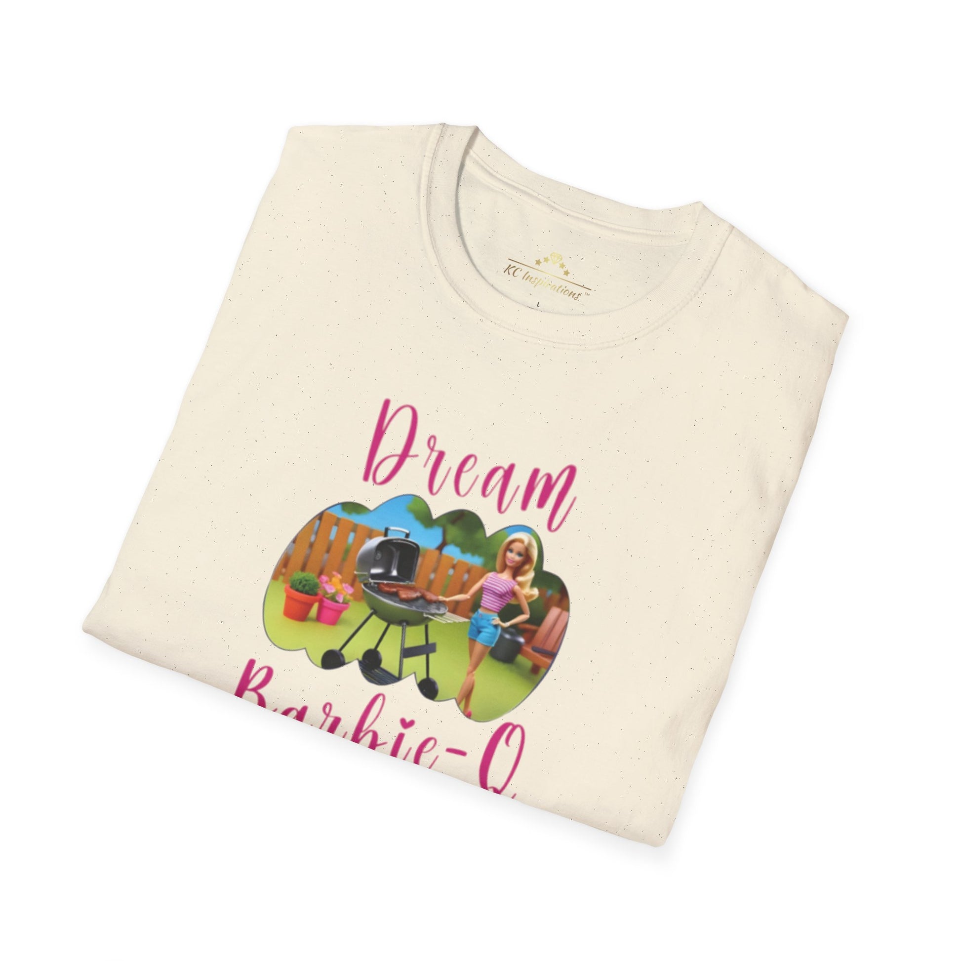 A beige T-shirt with speckles featuring a crisp graphic design of two dolls barbecuing in a backyard. The text "Dream Barbie-Q" is written in pink cursive above and below the image, making this Dream Barbie Q - K.C. BBQ Inspired T-Shirt by KC Inspirations perfect for fans of Kansas City BBQ.