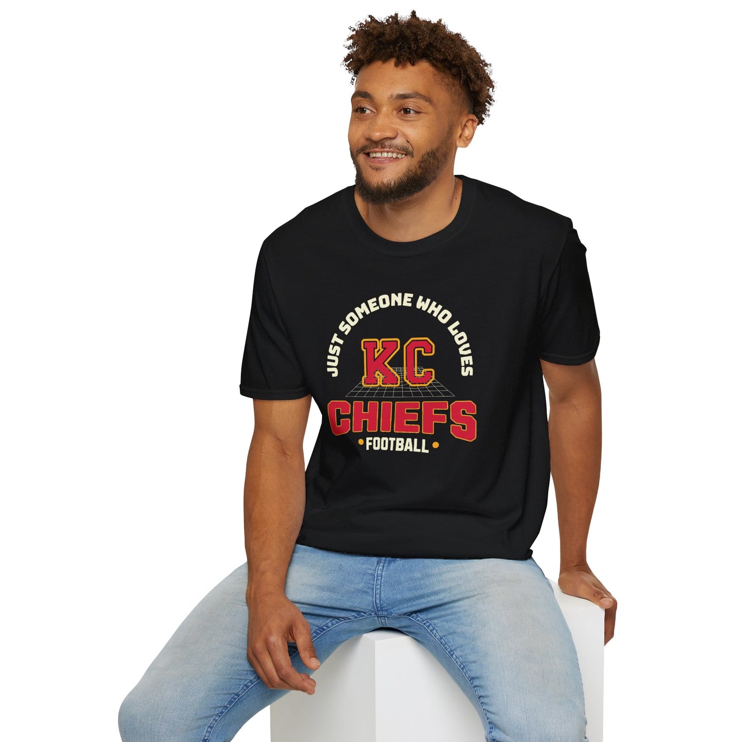 A smiling person with short curly hair and a beard is sitting on a white cube, wearing a black T-shirt that reads "Just someone who loves KC Chiefs football" in red and yellow text, showcasing true Chiefs pride, along with light blue jeans. The T-shirt is the Just Someone Who Loves - Kansas City Chiefs Inspired T-Shirt by KC Inspirations.