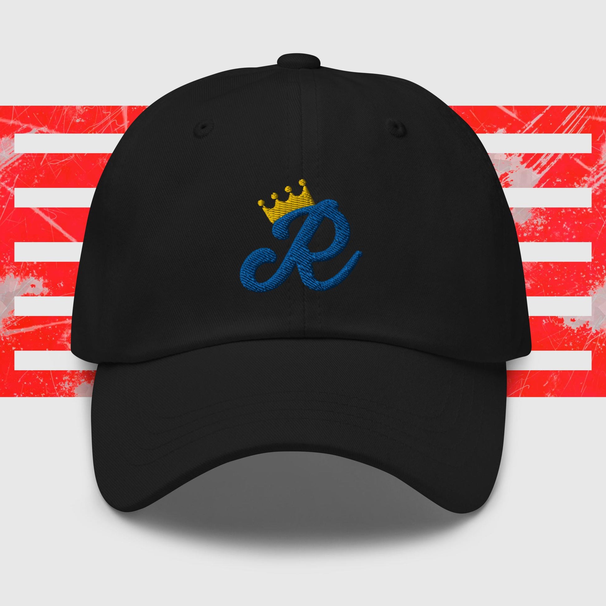 A white dad hat is centered in the image, featuring a blue cursive letter "R" embroidered on the front with a gold crown above it. The background consists of red and white horizontal stripes with a distressed texture, reminiscent of classic Kansas City Royals cap designs. This is the Royal Monogram - Kansas City Royals Inspired Cap from KC Inspirations.