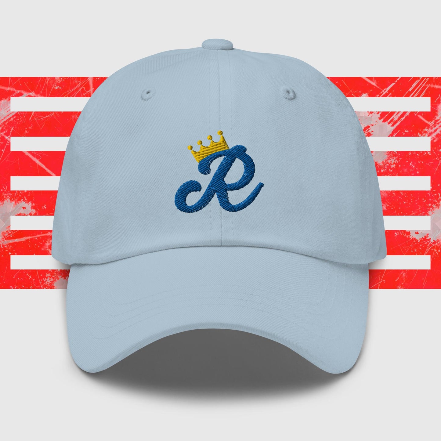 A white dad hat is centered in the image, featuring a blue cursive letter "R" embroidered on the front with a gold crown above it. The background consists of red and white horizontal stripes with a distressed texture, reminiscent of classic Kansas City Royals cap designs. This is the Royal Monogram - Kansas City Royals Inspired Cap from KC Inspirations.