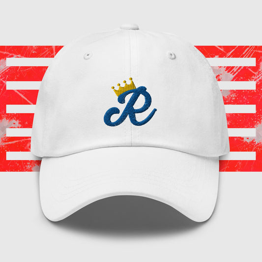 A white dad hat is centered in the image, featuring a blue cursive letter "R" embroidered on the front with a gold crown above it. The background consists of red and white horizontal stripes with a distressed texture, reminiscent of classic Kansas City Royals cap designs. This is the Royal Monogram - Kansas City Royals Inspired Cap from KC Inspirations.