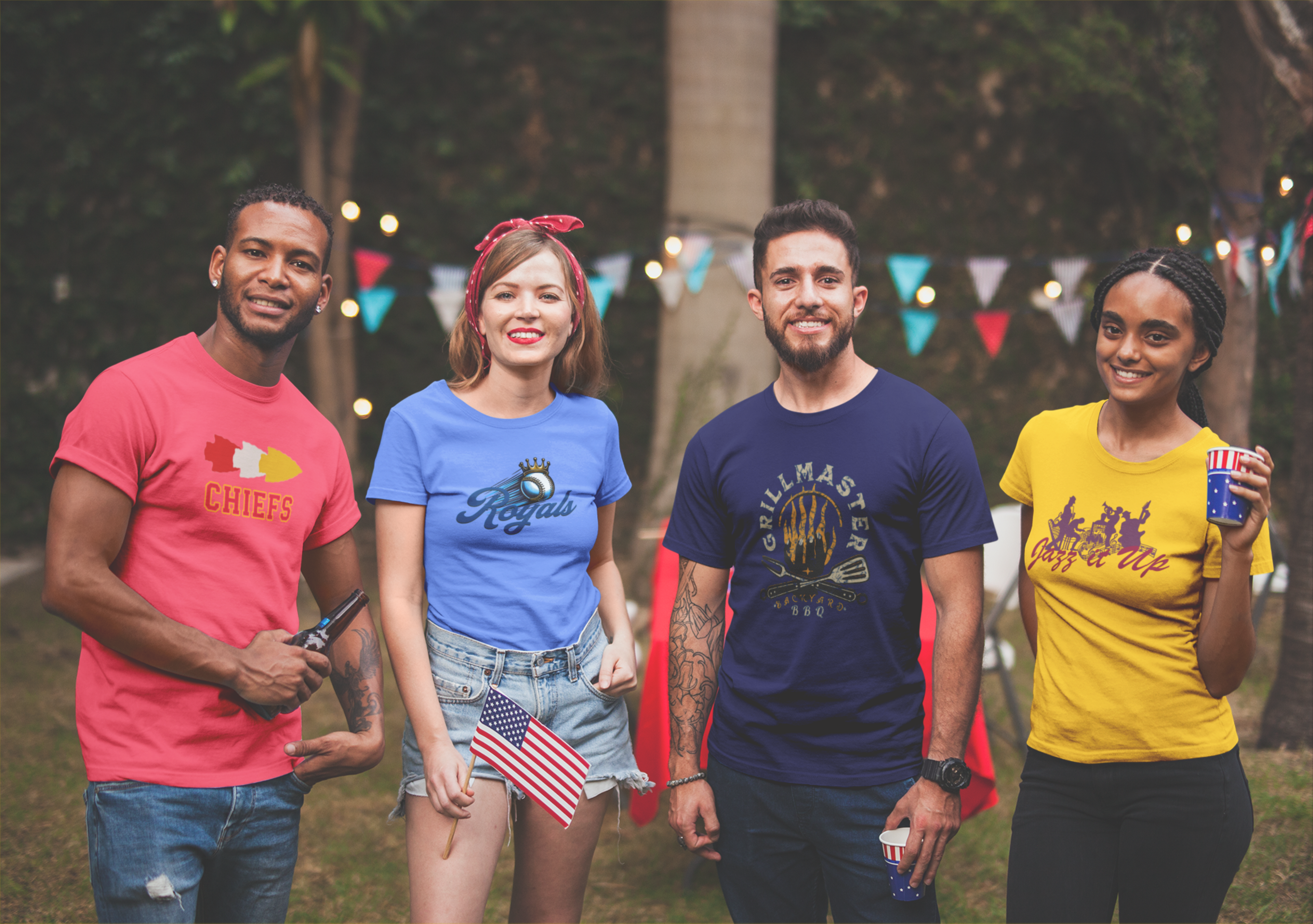 Four people stand outdoors at a festive KC Royals gathering, each wearing colorful t-shirts representing different Kansas City themes for the Chiefs the Royals KC Jazz and Grilling. String lights and triangular bunting decorate the background. They smile and hold drinks, one person also holding an American flag while grilling nearby.