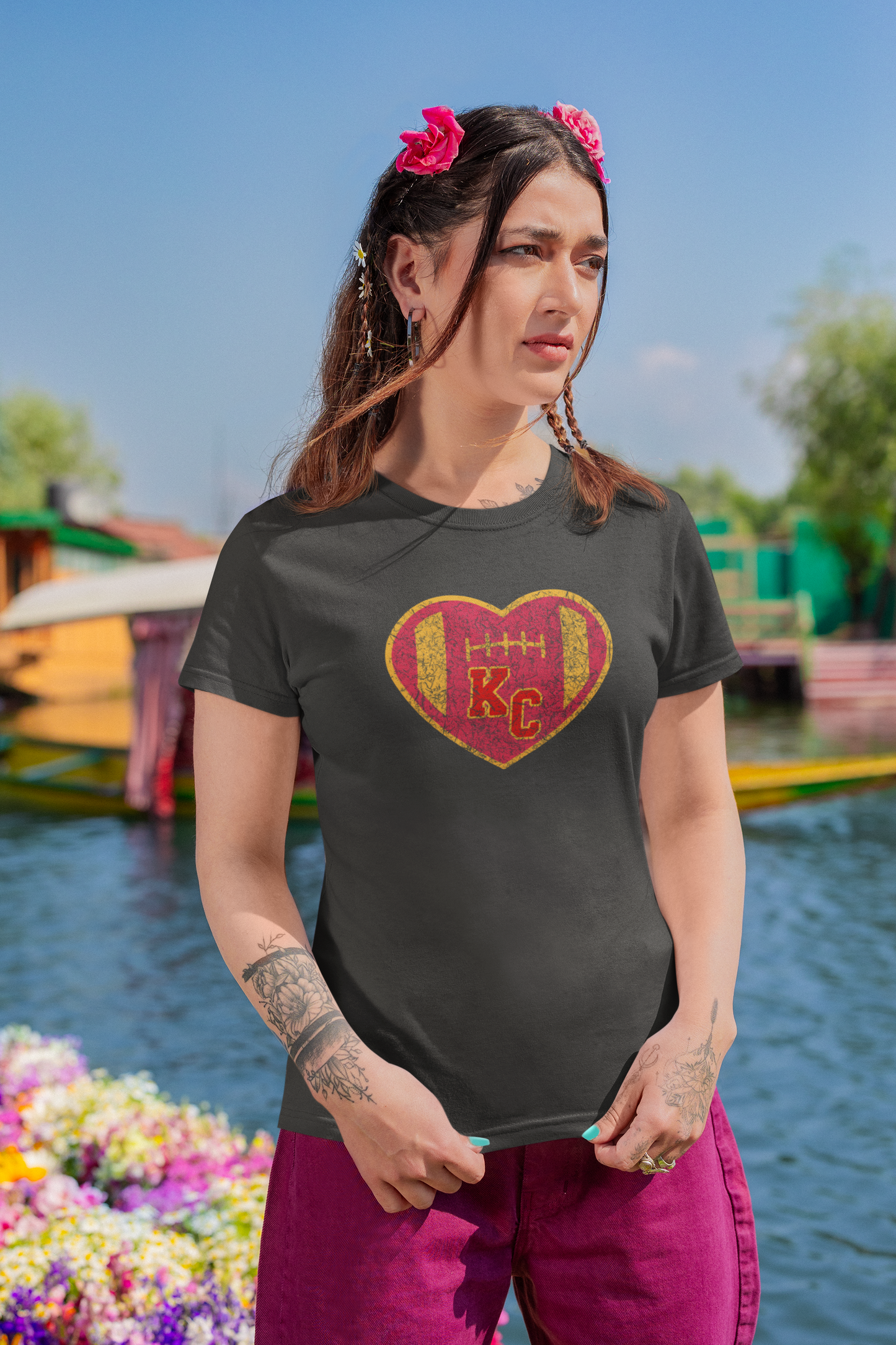 A dark gray women's T-shirt featuring a distressed heart-shaped design in the center. The heart, styled like a football with stitching details, is colored in red and gold. Inside the heart, the letters "KC" are prominently displayed in red with a yellow border. Perfect for Kansas City Chiefs fans! This is the Vintage KC Heart Red & Gold - Kansas City Chiefs Inspired Women's T-Shirt from KC Inspirations.